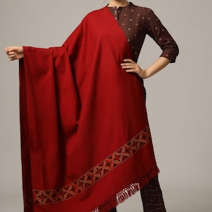 May include: A maroon shawl with a geometric patterned border and fringe. The shawl is draped over a person wearing a brown top with a geometric pattern.