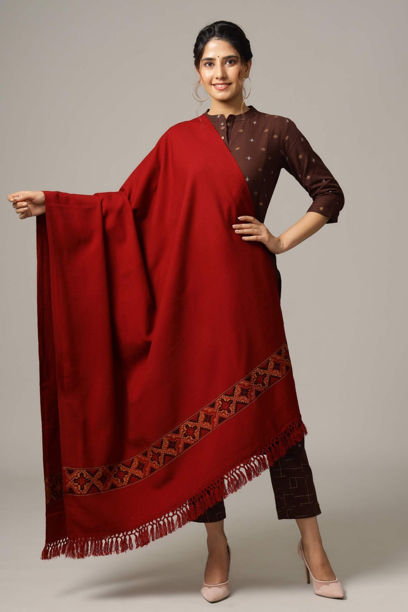 May include: A maroon shawl with a geometric patterned border and fringe. The shawl is draped over a person wearing a brown top with a geometric pattern.