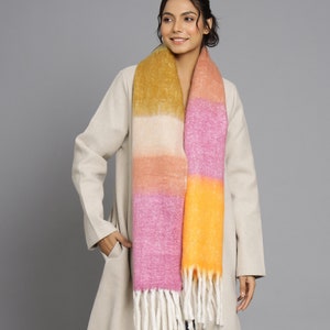 Puede incluir: Una bufanda a rayas con flecos. La bufanda es en tonos de amarillo, naranja, rosa y blanco.