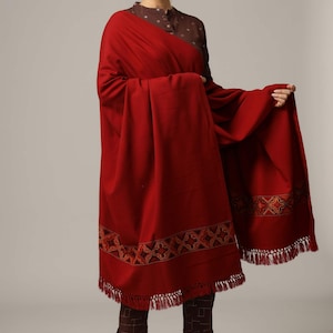 May include: A maroon shawl with a geometric embroidered border and fringe. The shawl is draped over a person wearing a brown top and pants.