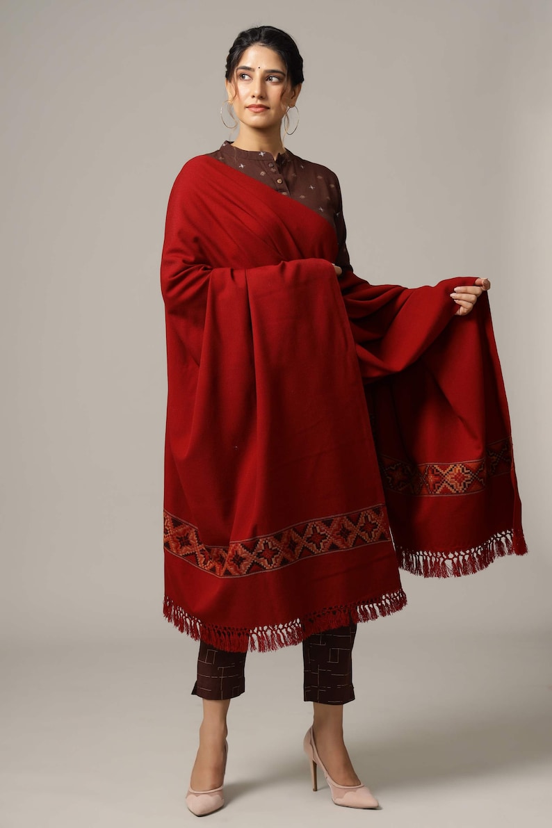 May include: A maroon shawl with a geometric embroidered border and fringe. The shawl is draped over a person wearing a brown top and pants.
