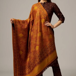 May include: A mustard yellow shawl with a geometric pattern and a maroon border. The shawl has a fringe trim and is made of a soft, luxurious fabric.