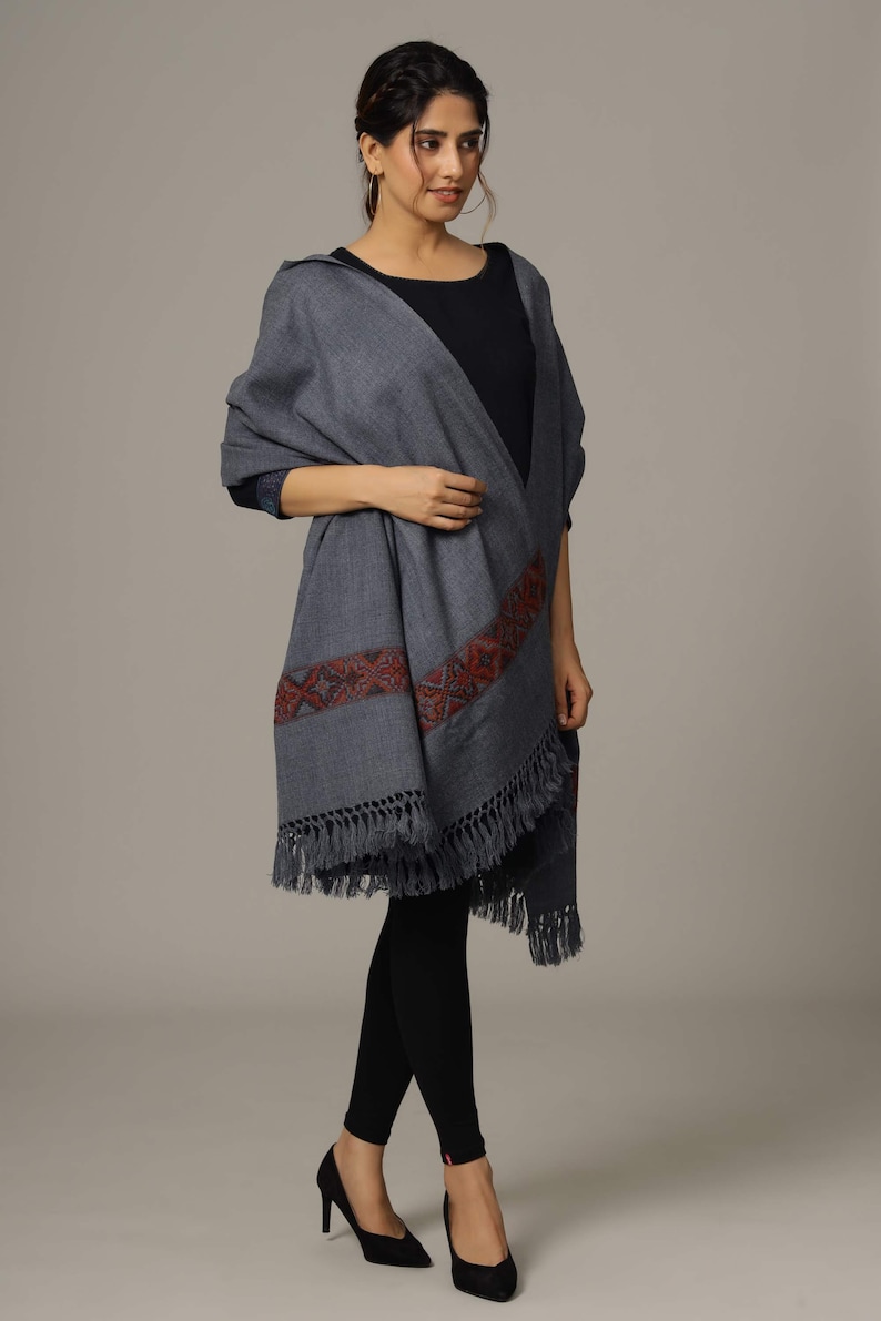 May include: A gray shawl with a red and blue embroidered border and fringe. The shawl is draped over a person's shoulders.