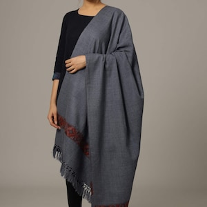 May include: A gray wool shawl with a red and blue embroidered border and fringe. The shawl is draped over a person's shoulders.