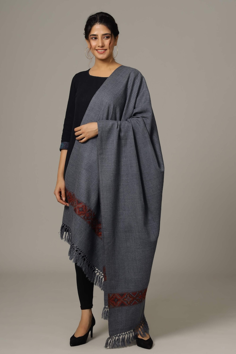 May include: A gray wool shawl with a red and blue embroidered border and fringe. The shawl is draped over a person's shoulders.