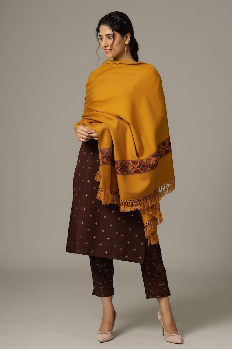 May include: A brown and white patterned kurta and pants set with a mustard yellow shawl featuring a colorful embroidered border and fringe. The shawl is draped over the shoulders and the model is wearing it with a casual, relaxed look.
