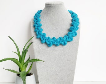 May include: A turquoise blue crocheted necklace with a knotted design. The necklace is displayed on a gray mannequin bust.