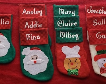 May include: Nine personalized Christmas stockings with different names embroidered on them. The stockings are red, green, and white with various holiday-themed appliques, including Santa Claus, a snowman, and a gingerbread man.