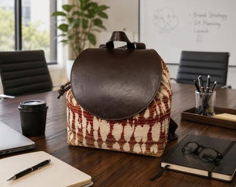 May include: A backpack featuring a large, smooth, dark brown leather flap closure and a main body made of a textured fabric with a repeating pattern of cream-colored vertical shapes against a deep red and brown background.