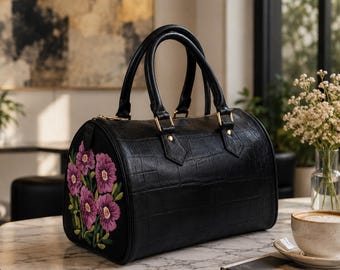 May include: A black barrel-shaped handbag with a crocodile-embossed texture and two top handles. The side panel features an embroidered design of purple flowers with green stems and leaves. The bag includes gold-toned hardware and a zipper closure.