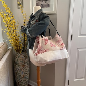 May include: A floral tote bag with a cream-colored base and a pink and red floral pattern. The bag is displayed on a mannequin wearing a denim jacket. The bag has black straps and a button closure.