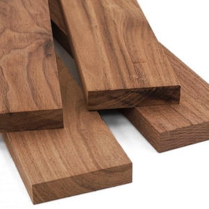 May include: Close-up of several rectangular, unfinished walnut wood planks. The planks are various sizes and arranged at different angles, showcasing the natural wood grain and rich brown color. The background is white.