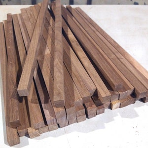 May include: A pile of wooden dowels, all the same size, with a square cross-section. The dowels are a light brown color and are arranged in a random pattern.