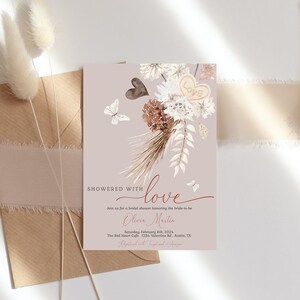 May include: A blush pink bridal shower invitation with a watercolor illustration of a bouquet of dried flowers, butterflies, and a heart with the word "love" on it. The text "Showered with love" is written in a script font. The invitation includes the bride-to-be's name, the date, time, and location of the shower.