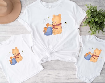 Puede incluir: Un conjunto de tres camisetas blancas con un personaje de dibujos animados de Winnie de Pooh sosteniendo un tarro de miel. Las camisetas son de diferentes tamaños, siendo la más grande la que tiene un diseño anudado en la parte inferior.
