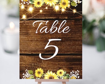 May include: Rustic wooden table number card with a floral design featuring sunflowers and white flowers. The card has a white background with the text "Table 5" in a cursive font.