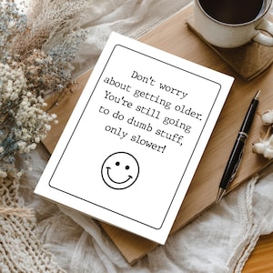 May include: A white rectangular greeting card with a black border featuring the text: Don't worry about getting older. You're still going to do dumb stuff, only slower! Below the text is a simple black line drawing of a smiling face emoji.