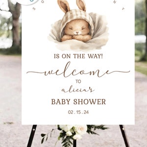 Pu&ograve; includere: Un cartello bianco con un'illustrazione di un coniglio marrone e il testo "IS ON THE WAY! welcome TO alicia's BABY SHOWER 02.15.24". Il cartello &egrave; su un cavalletto nero.