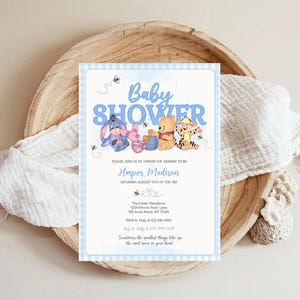 May include: Baby shower invitation with Winnie the Pooh characters. The invitation has a light blue gingham border and includes the text "Baby Shower," the name "Harper Madison," and event details. The invitation is on a wooden tray with a white cloth.