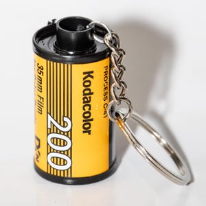 Puede incluir: Un llavero con un carrete de película Kodacolor 200 de 35 mm vintage. El carrete es amarillo con texto en blanco y negro, y una cadena y un llavero plateados están adjuntos. El texto incluye "Kodacolor", "200" y "35mm Film".
