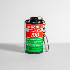 Puede incluir: Un llavero en miniatura de un carrete de película Fujicolor Super HR 100. El carrete tiene una etiqueta roja, blanca y verde con el texto "FUJICOLOR SUPER HR 100". El llavero incluye una cadena y un anillo plateados.