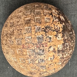 May include: A vintage golf ball with a faded white and black checkered pattern and the word "KRO-FLITE" in red lettering. The ball is worn and has a cracked surface.