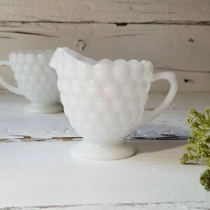 May include: Two white milk glass creamer pitchers with a textured, bubbled design. The pitchers have a handle and a round base.