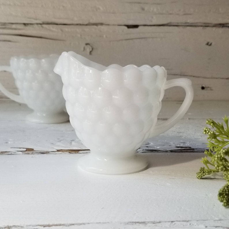 May include: Two white milk glass creamer pitchers with a textured, bubbled design. The pitchers have a handle and a round base.