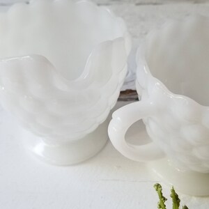 May include: Two white milk glass cups with a raised floral design. The cups have a scalloped rim and a footed base.