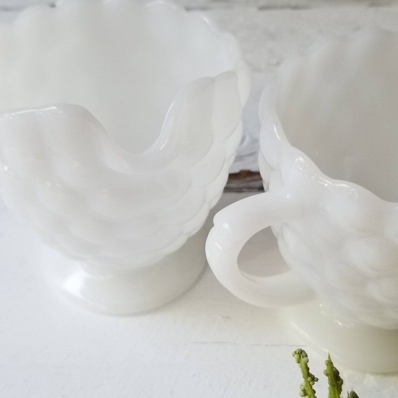 May include: Two white milk glass cups with a raised floral design. The cups have a scalloped rim and a footed base.