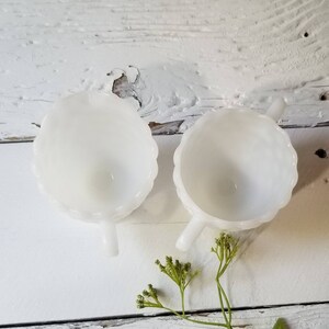 May include: Two white milk glass creamer cups with scalloped edges and handles. The cups are empty and sitting on a white wooden surface.