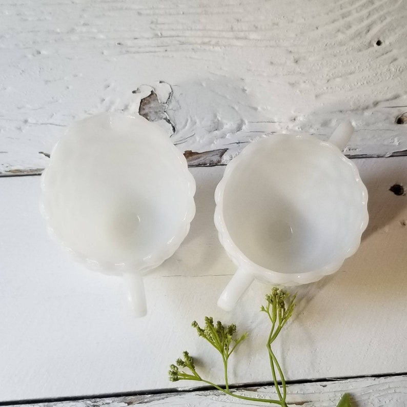 May include: Two white milk glass creamer cups with scalloped edges and handles. The cups are empty and sitting on a white wooden surface.