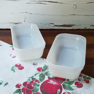 May include: Two white, rectangular, vintage glass baking dishes. The dishes are slightly angled and sit on a white tablecloth with a red cherry pattern. The background includes a distressed white wooden surface.