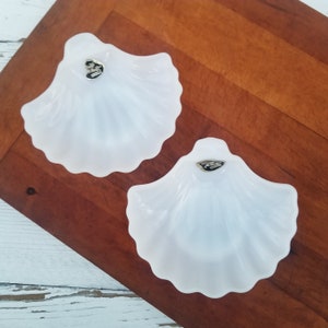 May include: Two white milk glass seashell-shaped dishes with gold trim. The dishes are on a wooden surface.