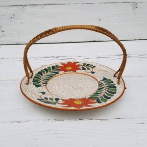 May include: A white ceramic plate with a woven wicker handle. The plate has a crackled glaze and is decorated with orange and green floral designs.