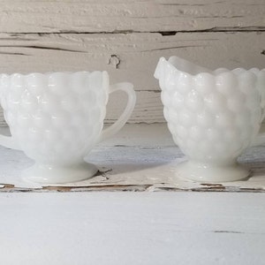 May include: A white milk glass creamer and sugar bowl set with a textured, bubbled design. The set is on a white wooden surface.