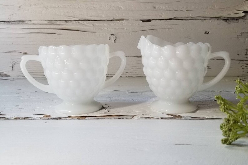May include: A white milk glass creamer and sugar bowl set with a textured, bubbled design. The set is on a white wooden surface.