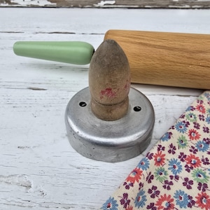 May include: A vintage metal cookie cutter with a wooden handle. The cutter is round and has a smooth, silver finish. The handle is brown and has red paint on it. The cutter is sitting on a white wooden surface.
