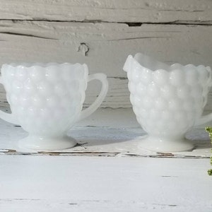 May include: A white milk glass creamer and sugar bowl set with a textured, bubble-like design. The set is on a white wooden surface.