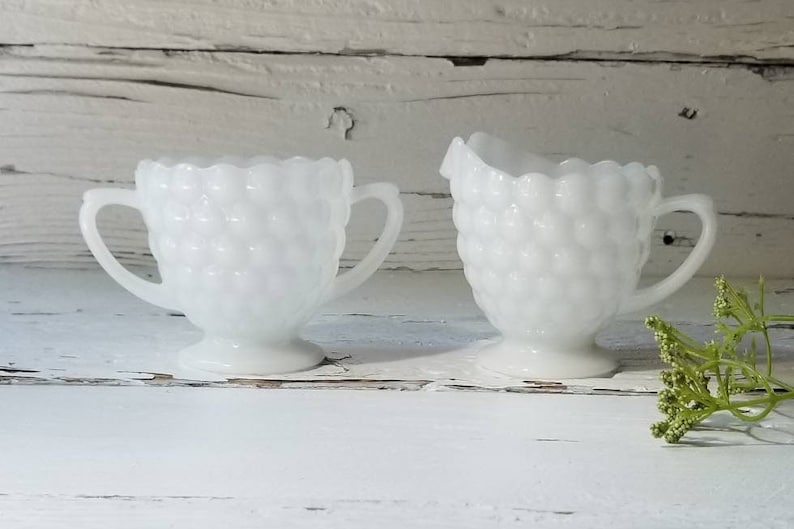May include: A white milk glass creamer and sugar bowl set with a textured, bubble-like design. The set is on a white wooden surface.