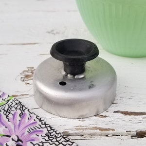 May include: A vintage metal donut cutter with a black handle. The cutter is circular with a small hole and is sitting on a white, distressed wooden surface. A green bowl is in the background.