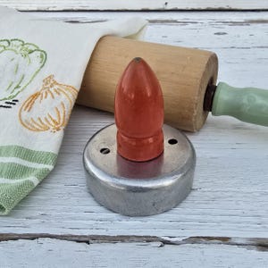 May include: A red wooden cookie cutter with a metal base. The cookie cutter is shaped like a bullet and has two small holes on the side. The cookie cutter is sitting on a white wooden surface.