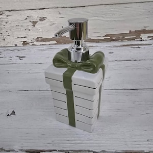 May include: White ceramic soap dispenser shaped like a gift box with a green ribbon and bow. The dispenser has a silver pump.