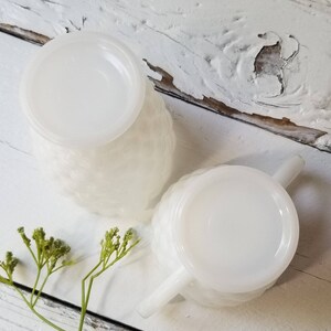 May include: Two white milk glass creamer pitchers with a textured design and handles. The pitchers are on a white wooden surface.