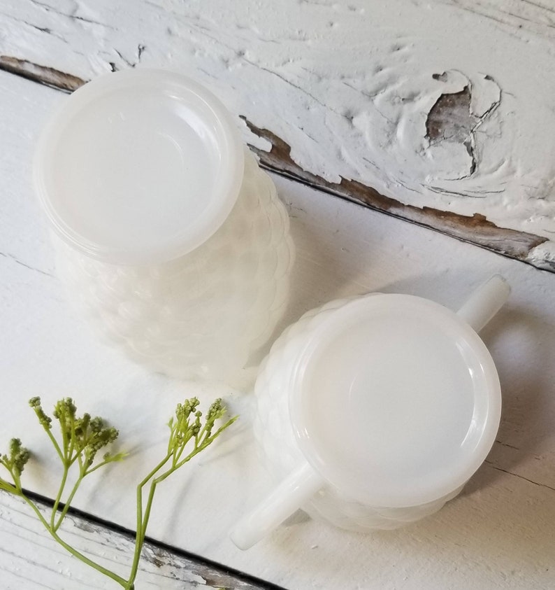 May include: Two white milk glass creamer pitchers with a textured design and handles. The pitchers are on a white wooden surface.
