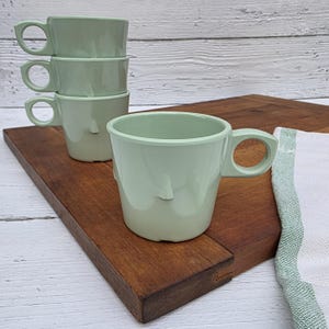 May include: Four light green ceramic mugs with unique handles are stacked on a wooden cutting board. One mug is positioned in front of the stack. A white and green striped towel is on the right side of the cutting board. The mugs have a minimalist design.