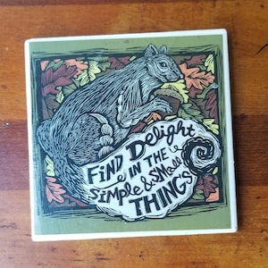 May include: Square ceramic coaster with a squirrel illustration. The squirrel is gray and holding an acorn, surrounded by fall leaves in shades of orange, red, and yellow. The coaster has the text "Find Delight in the Simple & Small Things."