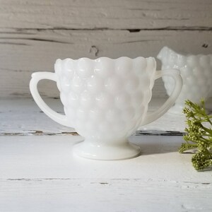 May include: White milk glass creamer with two handles and a textured surface. The creamer is on a white wooden surface.