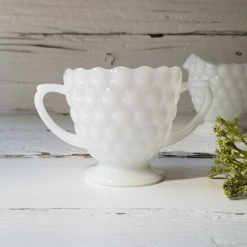 May include: White milk glass creamer with two handles and a textured surface. The creamer is on a white wooden surface.