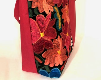 May include: A red tote bag with a black fabric panel featuring colorful embroidered flowers. The flowers are in shades of red, orange, yellow, blue, and green.